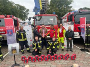 Feuerwehrleute des Landkreises Barnim stehen in Einsatzkleidung vor mehreren Feuerwehrautos. Vor ihnen liegen Defibrillatoren (AED-Geräte) und Übungspuppen zur Demonstration von Laienreanimation. Projektvorstellung ‚Herzsicher‘.