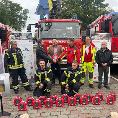 Feuerwehrleute des Landkreises Barnim stehen in Einsatzkleidung vor mehreren Feuerwehrautos. Vor ihnen liegen Defibrillatoren (AED-Geräte) und Übungspuppen zur Demonstration von Laienreanimation. Projektvorstellung ‚Herzsicher‘.